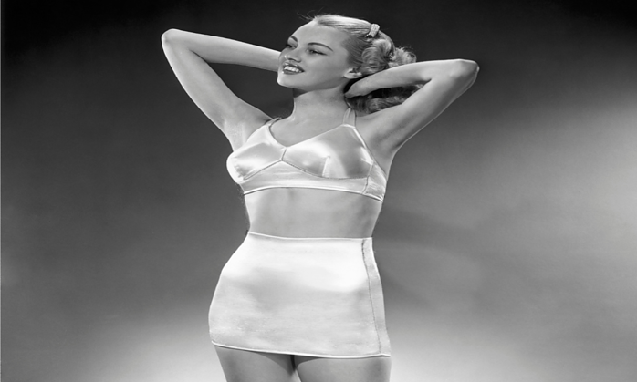 a black and white photo of a woman wearing retro shapewear from the 1950s smiling and hands above her head