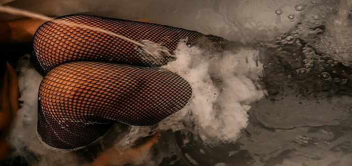 a woman wearing black fishnet bodysuit in a tub full of water, her knees bent poking out of the water