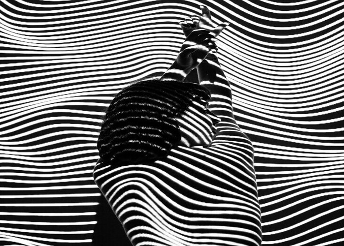 black and white artsy image of the backview of a woman from the waist up, head turned to the side and arms above her head. Black and white shadow stripes line the image horizontally