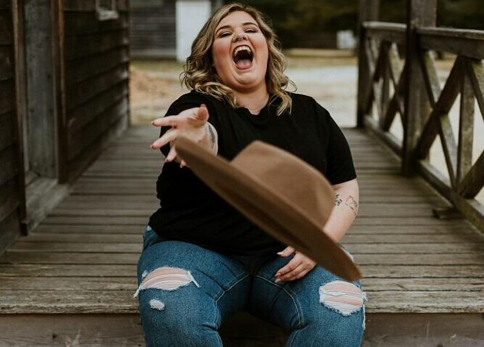 woman in jeans and t-shirt laughing and throwing a brown felt hat at the camera