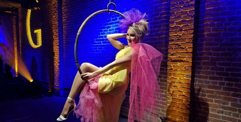 burlesque dancer sitting in a hanging hoop from the stage with one leg pointed out and a smile on her face