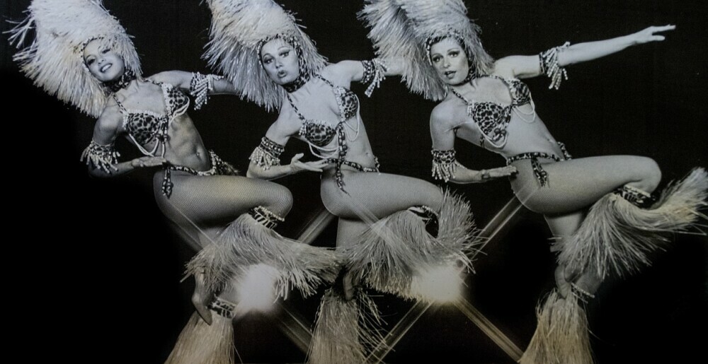 3-cabaret-dancers-with-one-leg-kicked-up-in-a-bend-on-stage-in-a-black-and-white-image